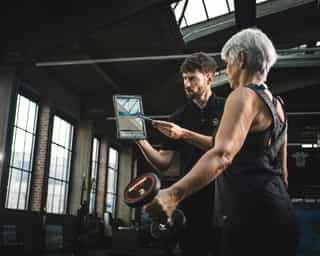 Trainer erläutert Patientin am Tablet die Kraftübungen mit Kurzhanteln.