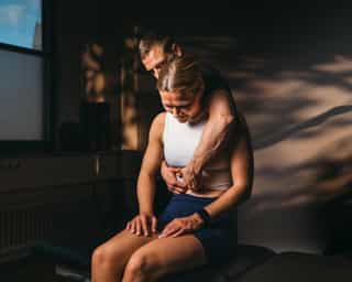 Patientin sitzt bei PhysioSport Köln, männlicher Therapeut führt osteopathische Behandlung am Rücken durch.