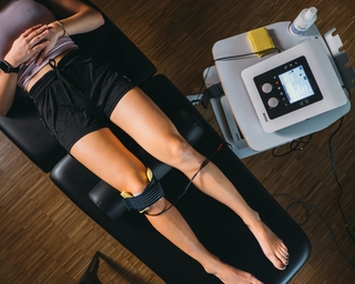 Patientin liegt auf Liege bei PhysioSport, Elektroden am Knie für Elektrotherapie in der Physikalischen Therapie, Gerät daneben.