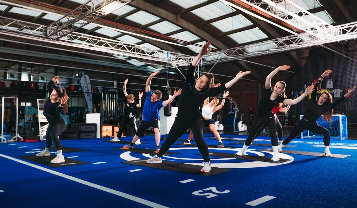 Zu sehen ist ein Gruppe von Patienten bei PhysioSport Köln, die gerade Gruppentraining in einer Trainingshalle mit blauem Boden durchführen.