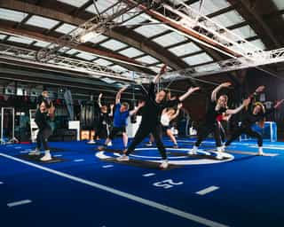 Zu sehen ist ein Gruppe von Patienten bei PhysioSport Köln, die gerade Gruppentraining in einer Trainingshalle mit blauem Boden durchführen.